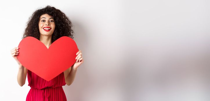 Romantic woman in dress showing big red heart, falling in love, smiling happy at camera, white background.