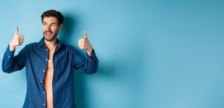 Handsome young man looking aside at empty space and showing thumbs up, standing on blue background.