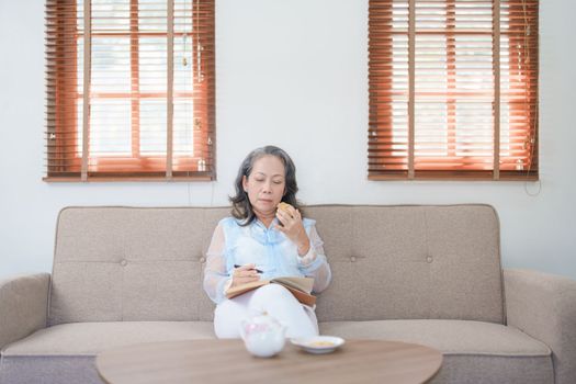 Portrait of an elderly Asian woman holding a notebook eating snacks and drinking tea