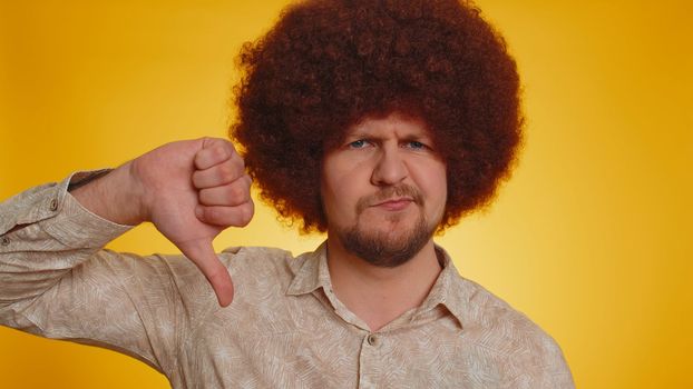 Dislike. Upset unhappy man with lush Afro hairstyle coiffure showing thumbs down sign gesture, expressing discontent, disapproval, dissatisfied, dislike. Young guy. Indoor studio on yellow background