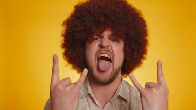 Rock n roll. Overjoyed delighted man with afro hairstyle showing gesture by hands, cool sign, shouting yeah with crazy expression, dancing, emotionally rejoicing in success win on yellow background