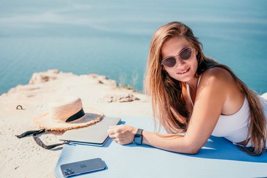 Successful business woman in yellow hat working on laptop by the sea. Pretty lady typing on computer at summer day outdoors. Freelance, travel and holidays concept.