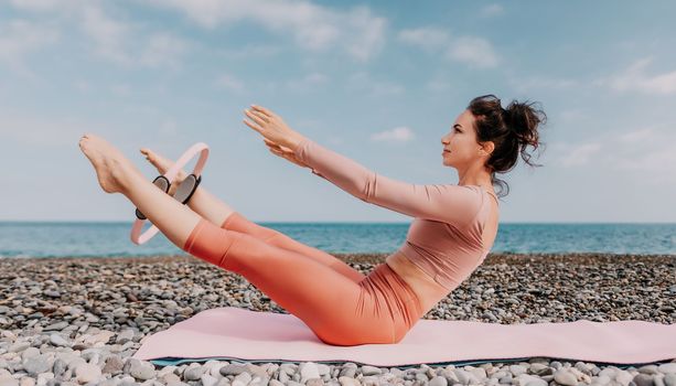 Middle aged well looking woman with black hair doing Pilates with the ring on the yoga mat near the sea on the pebble beach. Female fitness yoga concept. Healthy lifestyle, harmony and meditation.