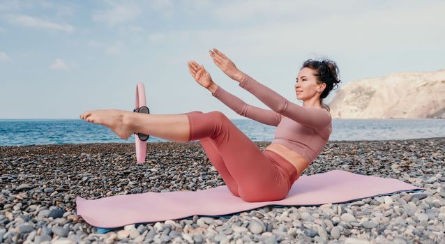 Middle aged well looking woman with black hair doing Pilates with the ring on the yoga mat near the sea on the pebble beach. Female fitness yoga concept. Healthy lifestyle, harmony and meditation.