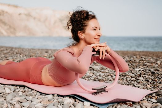 Middle aged well looking woman with black hair doing Pilates with the ring on the yoga mat near the sea on the pebble beach. Female fitness yoga concept. Healthy lifestyle, harmony and meditation.