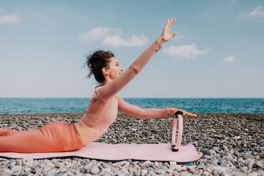 Middle aged well looking woman with black hair doing Pilates with the ring on the yoga mat near the sea on the pebble beach. Female fitness yoga concept. Healthy lifestyle, harmony and meditation.