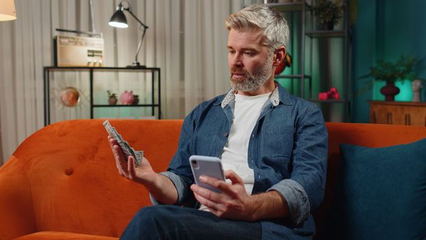 Planning family budget. Bearded senior man counting money dollar cash use smartphone calculate domestic bills at home. Joyful middle-aged guy satisfied of income saves money for planned vacation gifts