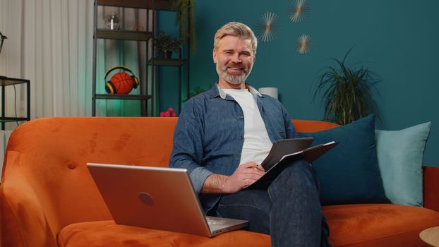 Middle-aged man accountant working with financial graph documents, calculate taxes for charts and graph reports of home income on couch. Businessman working on laptop computer planning expenses budget