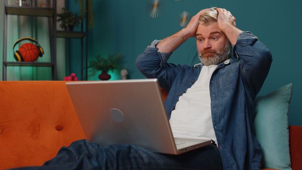 Displeased middle-aged bearded man use laptop typing browsing working, loses becoming surprised sudden lottery results bad news, fortune loss fail. Adult mature guy at home in evening room on couch