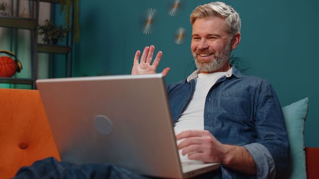 Middle-aged senior man sitting on couch looking at camera, making video webcam conference call with friends or family, enjoying pleasant conversation. Mature guy laughing waving hello at night home
