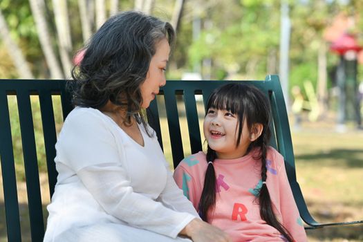 Attentive elderly grandmother and little grandchild having casual conversation, while sitting on a bench in the park.