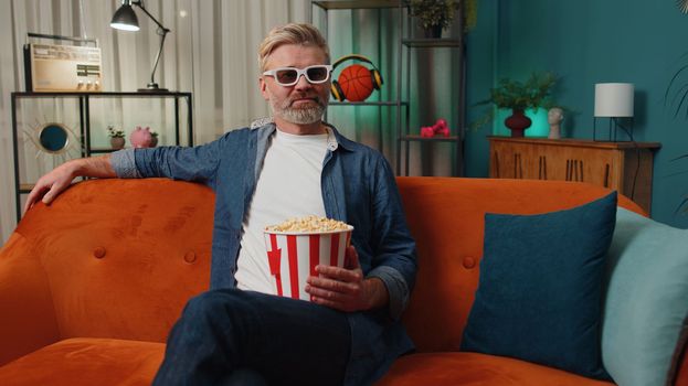 Excited bearded middle-aged man sits on sofa eating popcorn snacks and watching interesting TV serial, sport game, film, online social media movie content at home. Guy enjoying evening entertainment