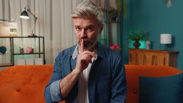 Shh be quiet, hush please. Bearded middle-aged senior man presses index finger to lips makes silence hush gesture sign do not tells gossip secret. Mature old guy at home evening room sitting on couch