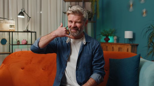Call me, here is contact number. Middle-aged man looking at camera advertising doing phone gesture like says hey you call me back at home. Mature senior gray-haired old guy at evening room apartment