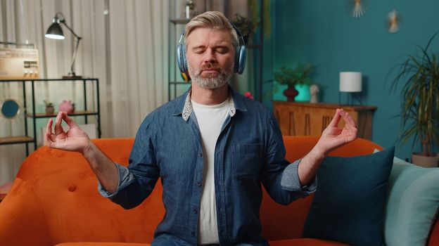 Keep calm down, relax. Middle-aged old man at home couch breathes deeply with mudra gesture, eyes closed meditating with concentrated thoughts, peaceful mind, listen music. Mature guy in evening room