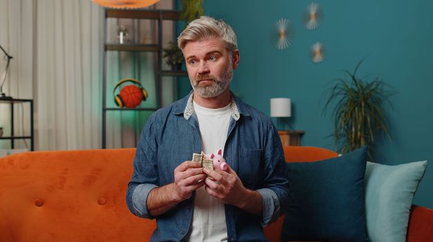 Poor middle-aged man insufficient amount of money, holding piggybank and one dollar banknote at home room. Financial crisis. Bankruptcy. Poverty and destitution at home. Mature senior gray-haired guy