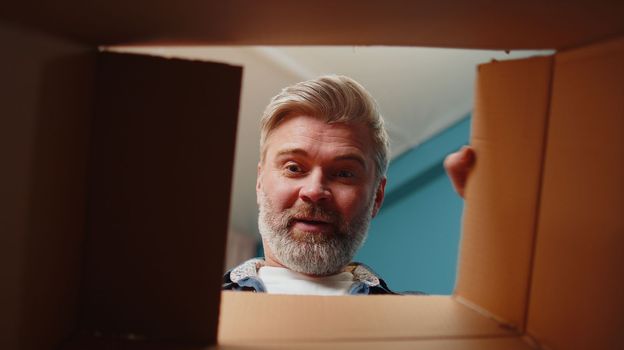 Happy surprised winner man unpacking delivery parcel at home. Smiling satisfied senior guy shopper online shop customer opening cardboard box receiving purchase gift headphones by fast postal shipping