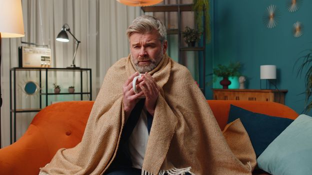 Sick man wrapped in plaid sit alone shivering from cold on sofa drinking hot tea in unheated apartment without heating due debt. Unhealthy senior guy feeling discomfort try to warm up at evening home