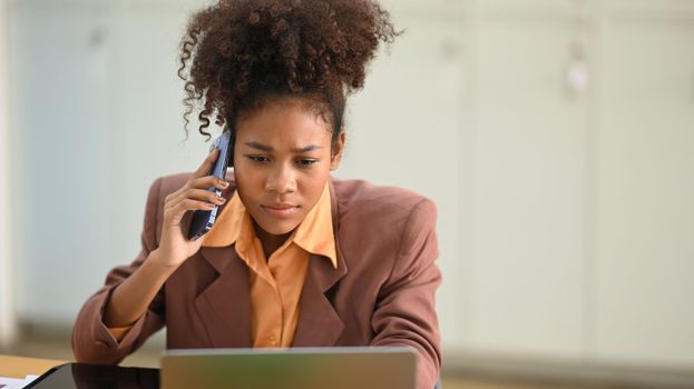 Young serious African office worker looking at laptop screen and talking on mobile phone, arguing with a client or customer.