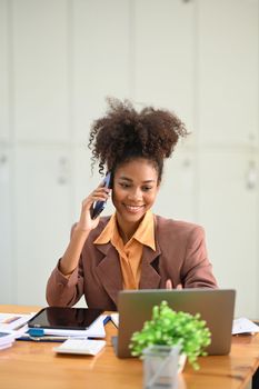 Positive American African female office worker having pleasant conversation on mobile phone and using laptop in office.