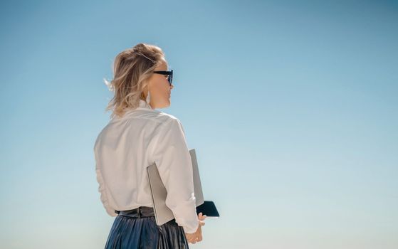 Business woman on nature in white shirt and black skirt. She works with an iPad in the open air with a beautiful view of the sea. The concept of remote work