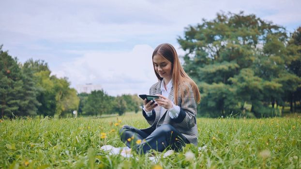 A young girl makes a purchase with her phone and bank card in the park, sitting on the grass