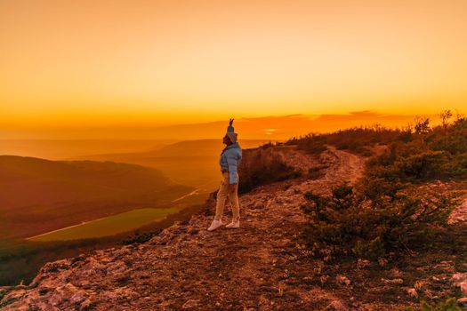 Woman tourist on top of sunrise mountain. The girl salutes the sun, wearing a blue jacket, white hat and white jeans. Conceptual design