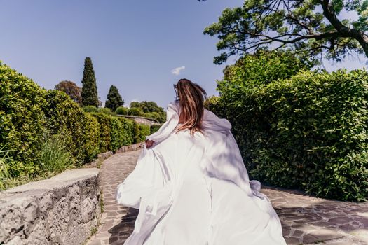 Brunette runs white dress park. A beautiful woman with long brown hair and a long white dress runs along the path along the beautiful bushes in the park, rear view.