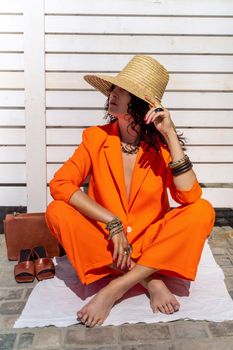 Stylish woman in an orange suit with a hat sits on a rug on a white striped background. On the hands are jewelry rings and bracelets, sandals and a bag stand side by side