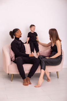 Happy multiracial family with little boy, in black clothes, with bare foot , sits in the light living room on light pink sofa. Vertical. Copy space