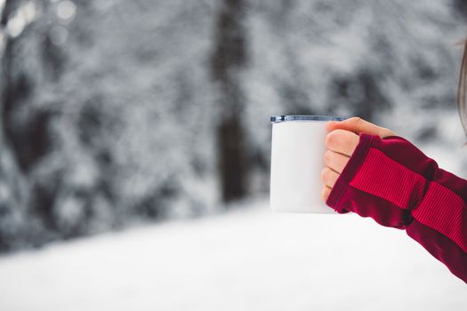 Beautiful young caucasian woman with brown hair, wearing a red jacket and a hat outside in the forest when it's snowing. Woman walking around a snowy forest. Woman holding a to go cup with tea.