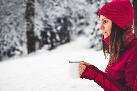 Beautiful young caucasian woman with brown hair, wearing a red jacket and a hat outside in the forest when it's snowing. Woman walking around a snowy forest. Woman holding a to go cup with tea.
