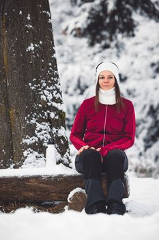 Beautiful young caucasian woman with brown hair, wearing a red jacket and a hat outside in the forest when it's snowing. Woman walking around a snowy forest. Woman holding a to go cup with tea.