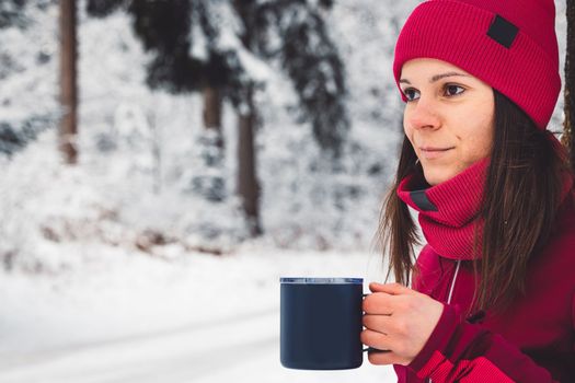 Beautiful young caucasian woman with brown hair, wearing a red jacket and a hat outside in the forest when it's snowing. Woman walking around a snowy forest. Woman holding a to go cup with tea.