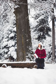 Beautiful young caucasian woman with brown hair, wearing a red jacket and a hat outside in the forest when it's snowing. Woman walking around a snowy forest. Woman holding a to go cup with tea.