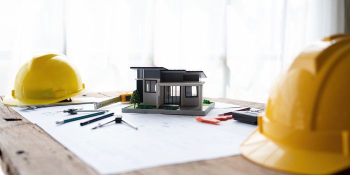 architectural model of houses on desk with drawing technical tools safty helmet and blueprint rolls for building construction plan, interior designer and architect work concept.