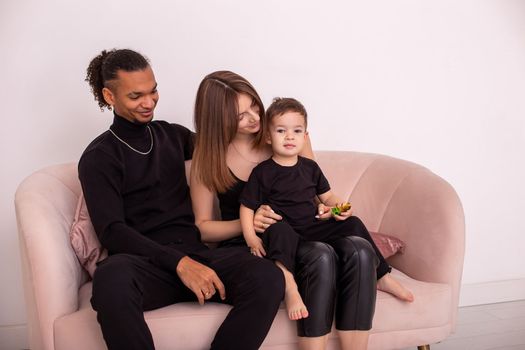 A beautiful young family with a young son, in black clothes, sits in the light living room on light pink sofa, by wall. Copy space. Close up