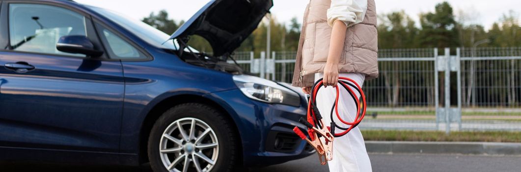 woman close-up with wires for charging the battery near the broken car with the hood up.