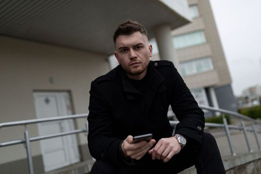 young man with a phone in his hands sitting looking at the camera outside.