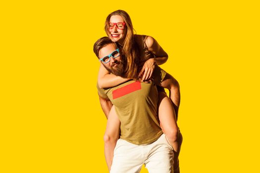 Portrait of happy joyful cheerful young couple looking at camera, man holding his girlfriend on his back, expressing positive emotions. Indoor studio shot isolated on yellow background.