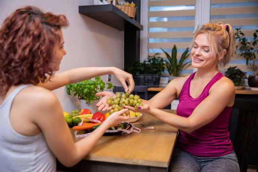 Close-up on blurred profile of woman with curly hair. A second woman sits across from her. Female colleagues serve themselves green grapes over the kitchen table.