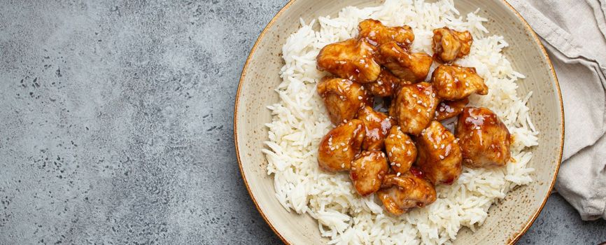 Chinese sweet and sour sticky chicken with sesame seeds and rice on ceramic plate with chopsticks top view on gray rustic stone background, traditional dish of China. Space for text