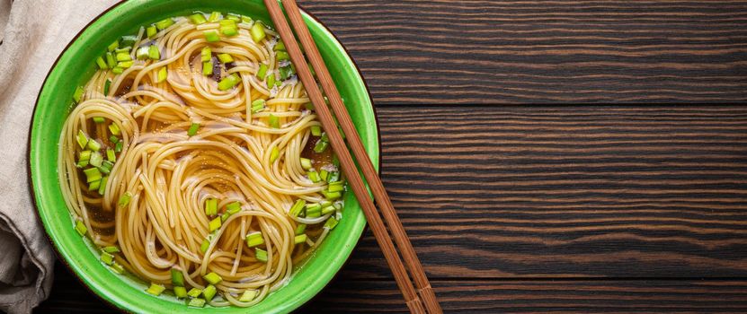 Asian noodles soup in green rustic ceramic bowl with wooden chopsticks top view on dark wooden background. Lo mein noodles with bouillon and green onion, space for text