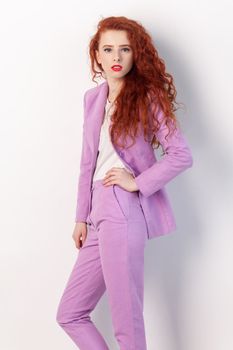 Portrait of attractive adorable confident ginger woman with curly hair, standing keeping hand on hip, looking at camera, wearing lilac suit. Indoor studio shot isolated on gray background.