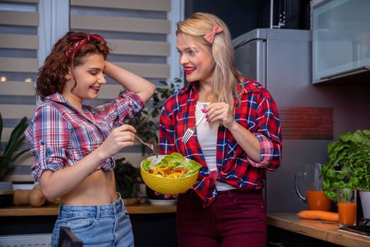 Two women are talking while standing in the kitchen eating a salad with peppers from one bowl. On the counter in the background stands a large pitcher of orange juice and next to it a glass. In the background is a curtained window.