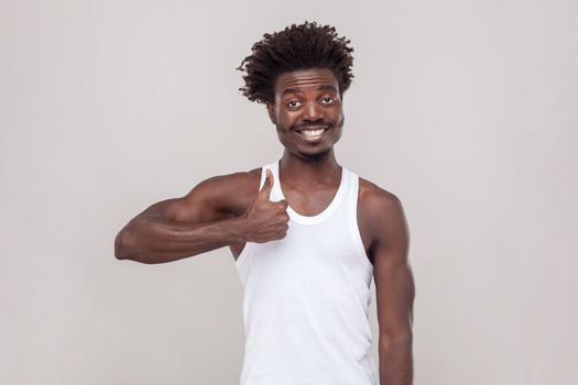 Nice joke. Man with Afro hairstyle showing like gesture, satisfied with something, being in good mood, shows her agreement, wearing white T-shirt. Indoor studio shot isolated on gray background.