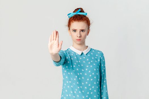 Portrait of strict bossy red haired woman with bun hairstyle, standing standing showing stop gesture with palm, looking at camera, wearing blue dress. Indoor studio shot isolated on gray background.