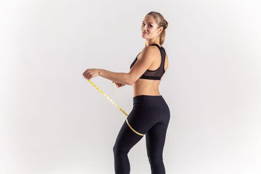 Side view portrait of delighted sporty blonde woman holding centimeter on her leg, checking result after sport marayhon, wearing black fitness clothing. Indoor studio shot isolated on gray background.