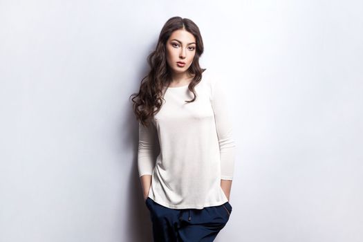 Portrait of calm serious woman with wavy hair wearing white shirt standing with hands in pockets, looking at camera with strict confident expression. Indoor studio shot isolated on gray background.