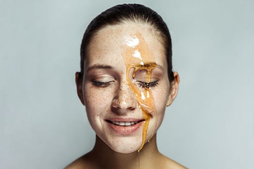 Portrait of happy beautiful young brunette woman with freckles and honey on face with closed eyes, posing toothy smile and happy face. Indoor studio shot isolated on gray background.
